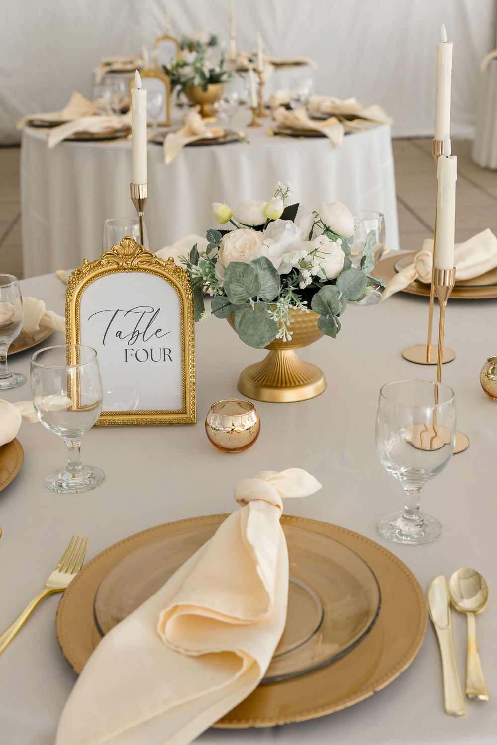 Elegant wedding table setting with gold charger, cream napkin, table four sign, white floral centerpiece and taper candles.
