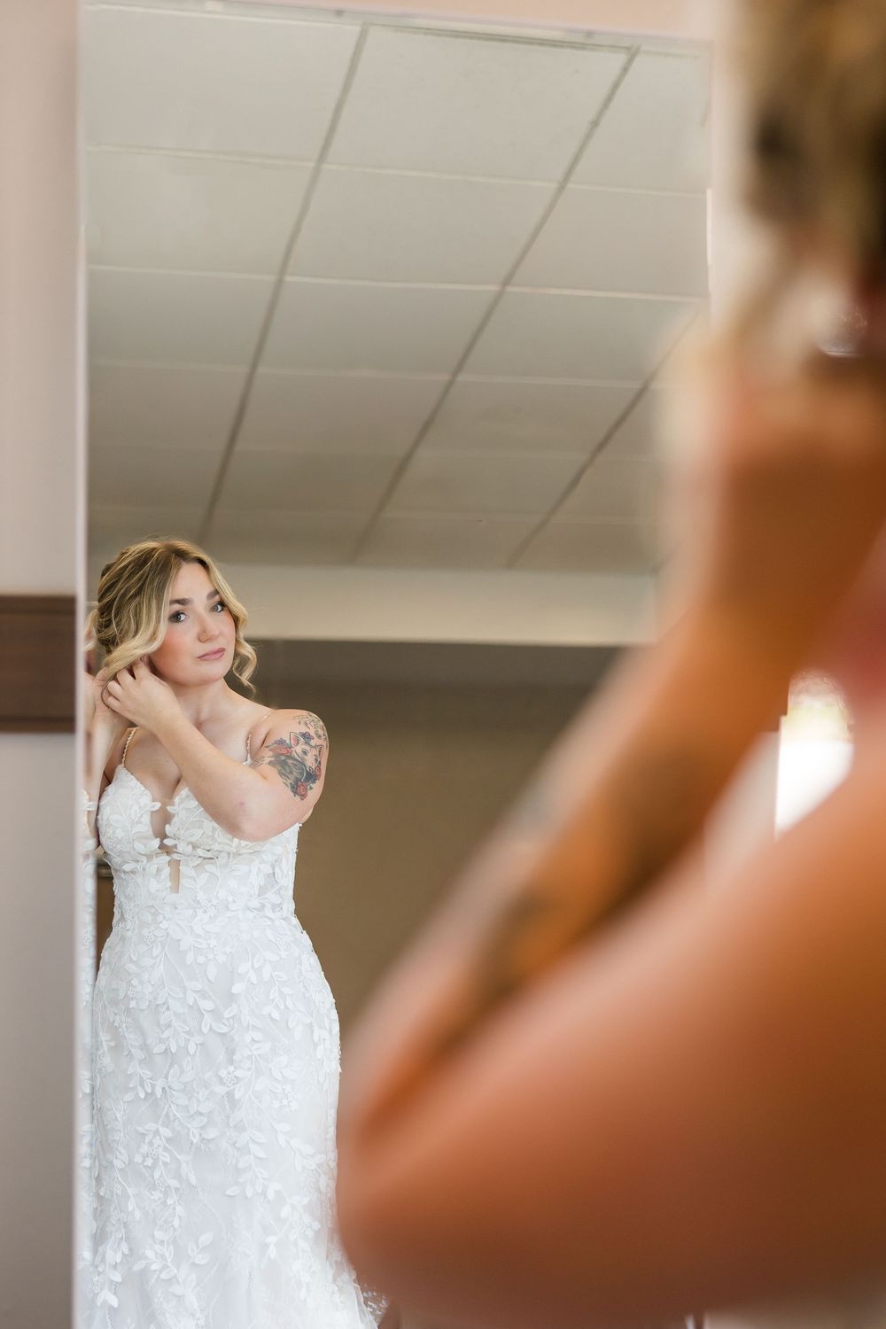 Bride in a white lace wedding gown with a shoulder tattoo adjusts her hair while looking into a mirror.