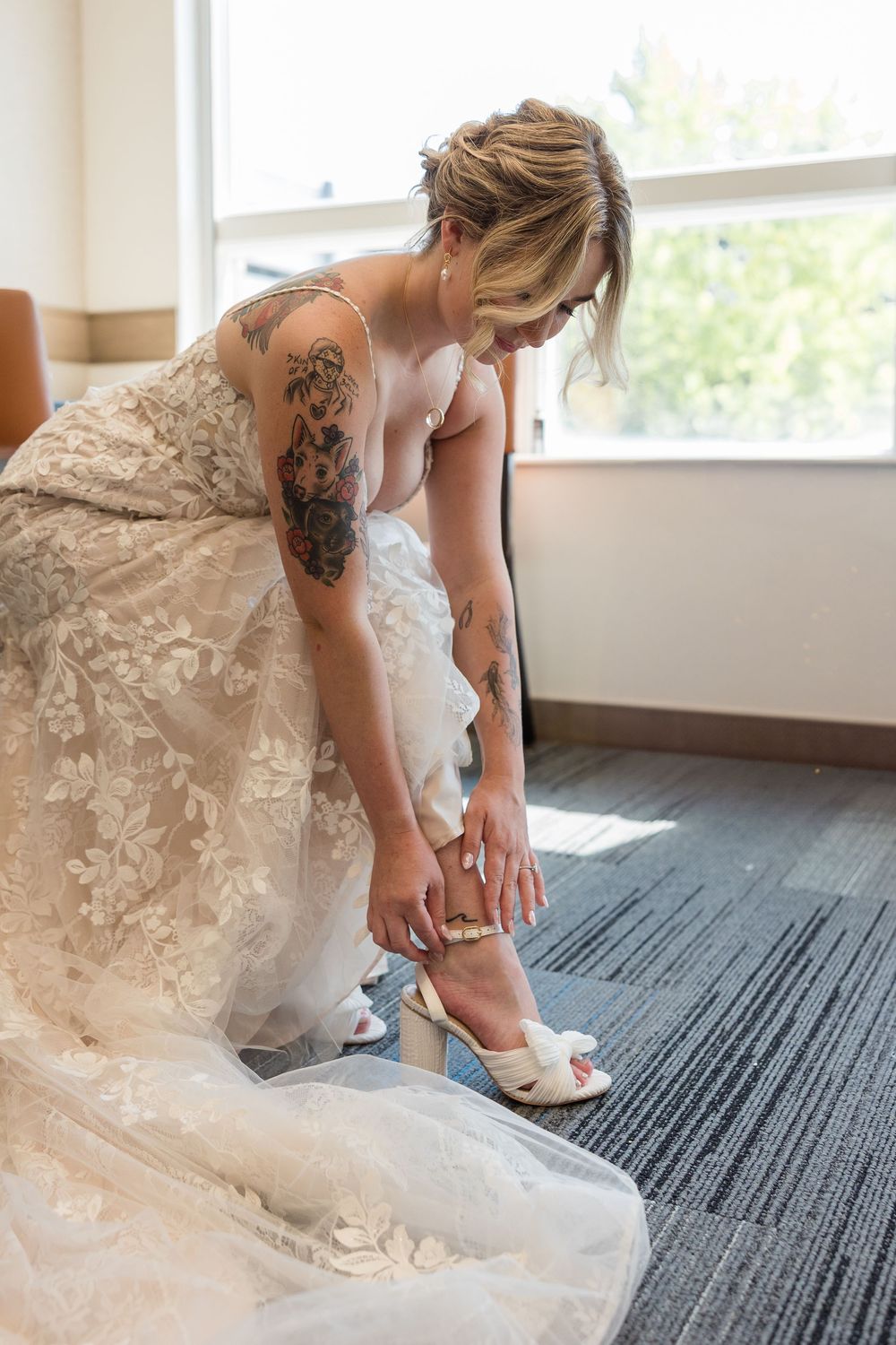 Tattooed bride in a floral lace wedding dress bends to fasten her white bow-detailed heels beside a sunny window.