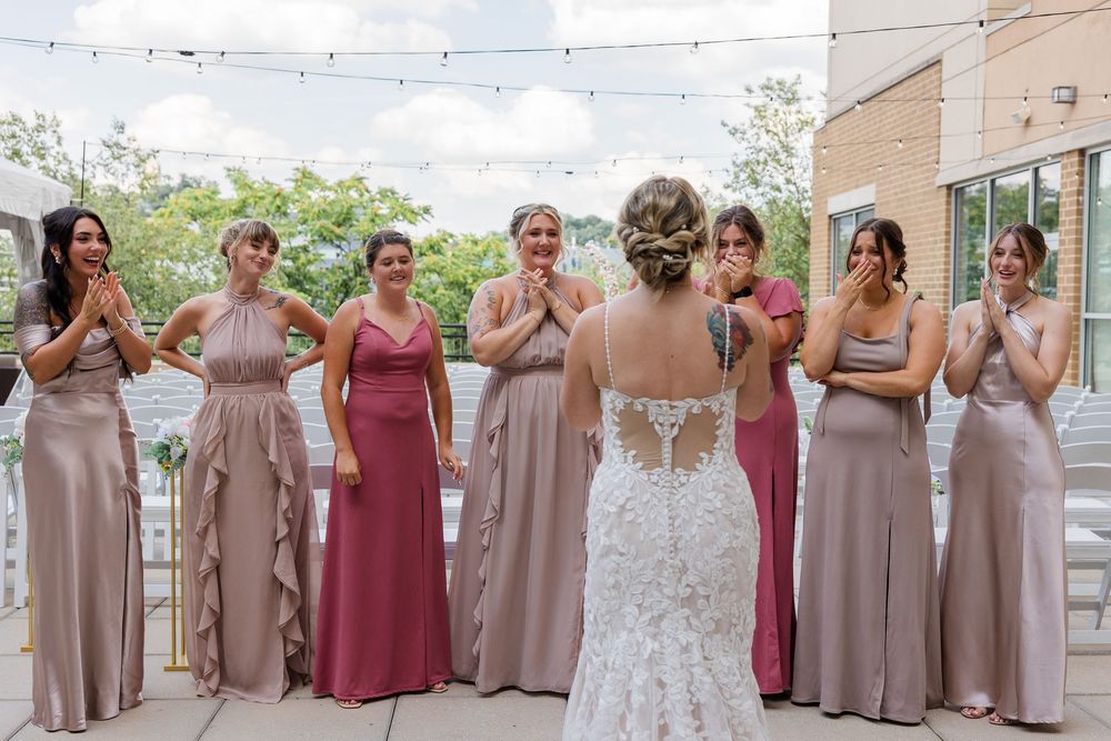 Bride in a lace wedding gown with an updo faces smiling bridesmaids in mauve and rose dresses during an outdoor wedding reveal.