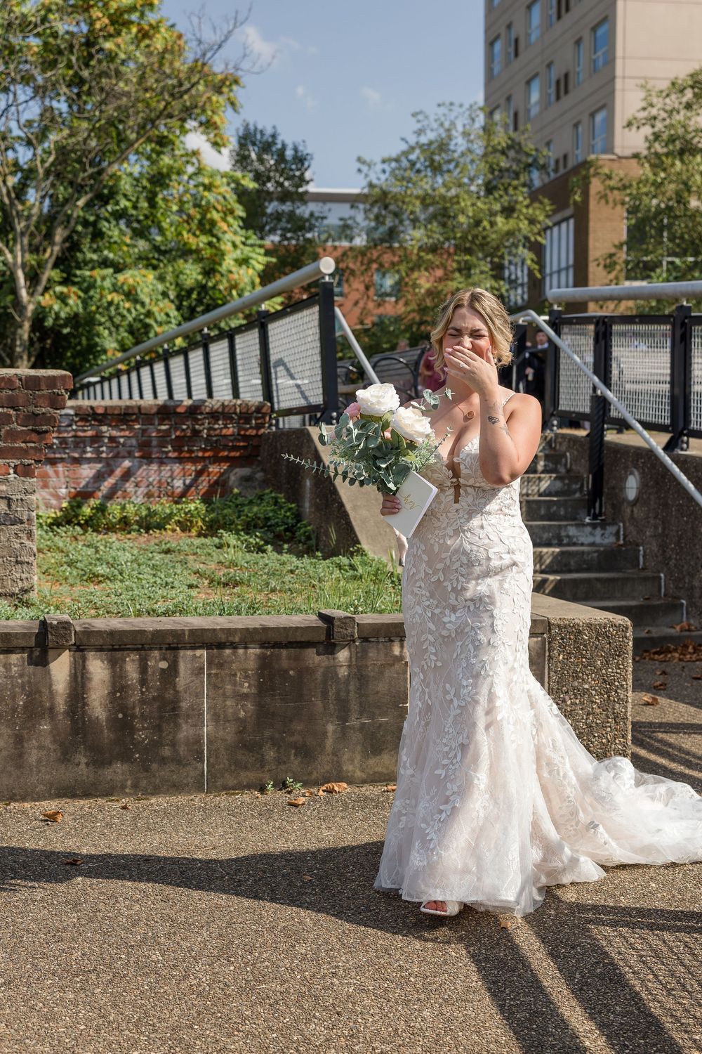 Emotional bride in a lace wedding gown holds a bouquet and covers her mouth with her hand beside a brick wall and outdoor staircase.