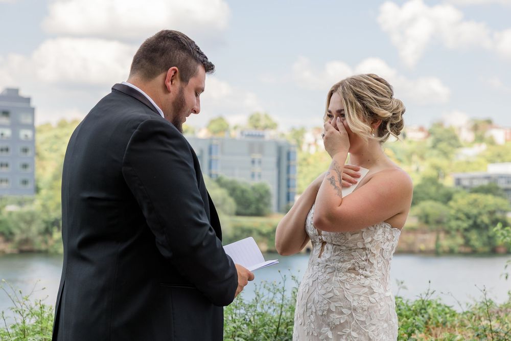 Emotional bride in a lace wedding gown wipes her tears as the groom in a dark suit reads his vows beside a scenic waterfront.