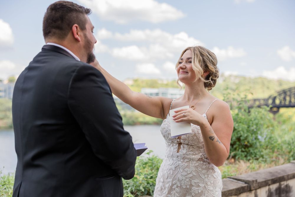 A bride in a lace wedding gown smiles and touches the groom's face during an outdoor ceremony with a river and bridge behind them.