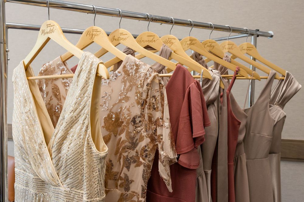 Wooden hangers hold elegant dresses in cream sequins, rose lace, terracotta, and taupe satin on a metal clothing rack.