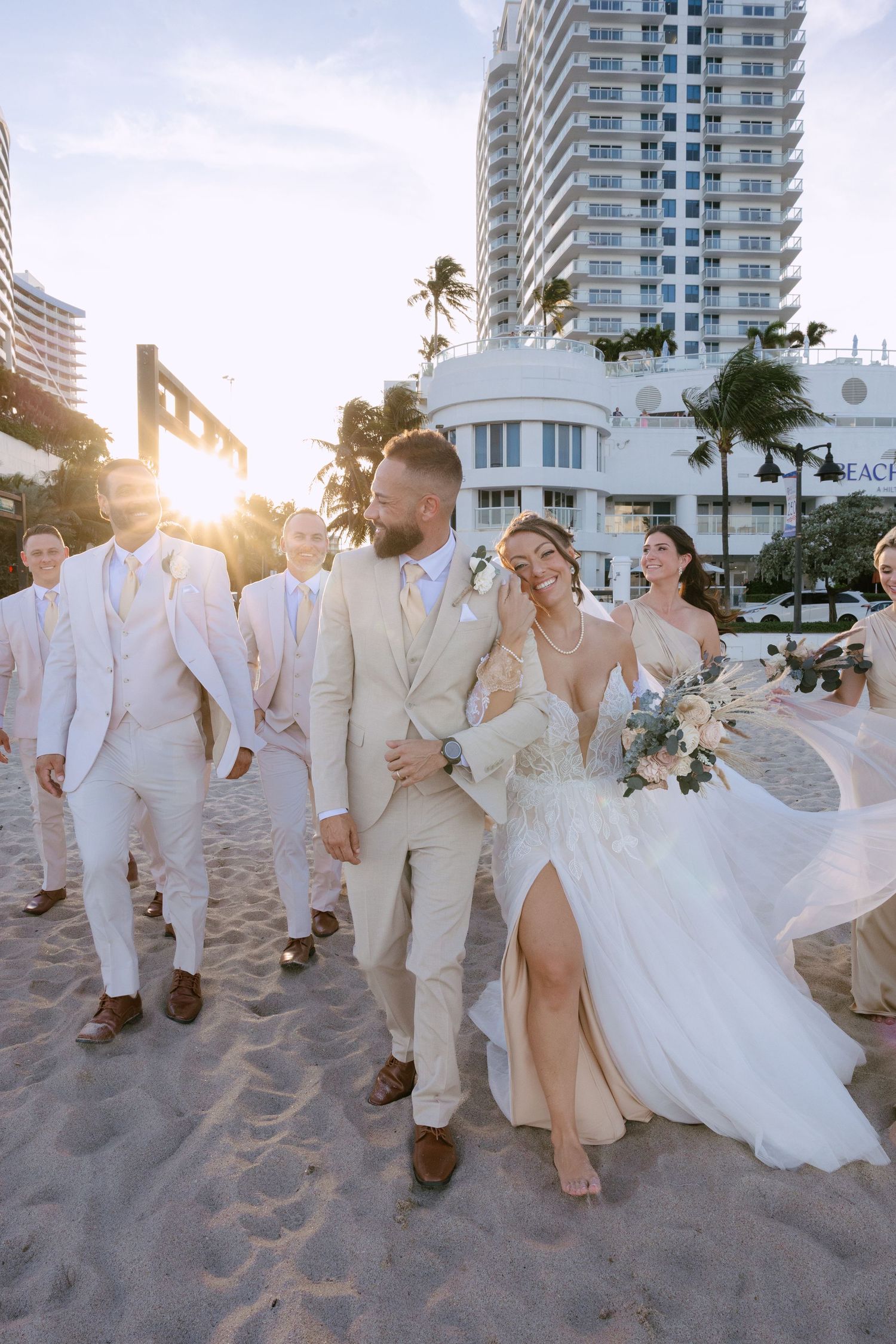 Bride in a flowing white gown and groom in a cream suit walk with groomsmen along a sunny beach wedding at Fort Lauderdale Beach