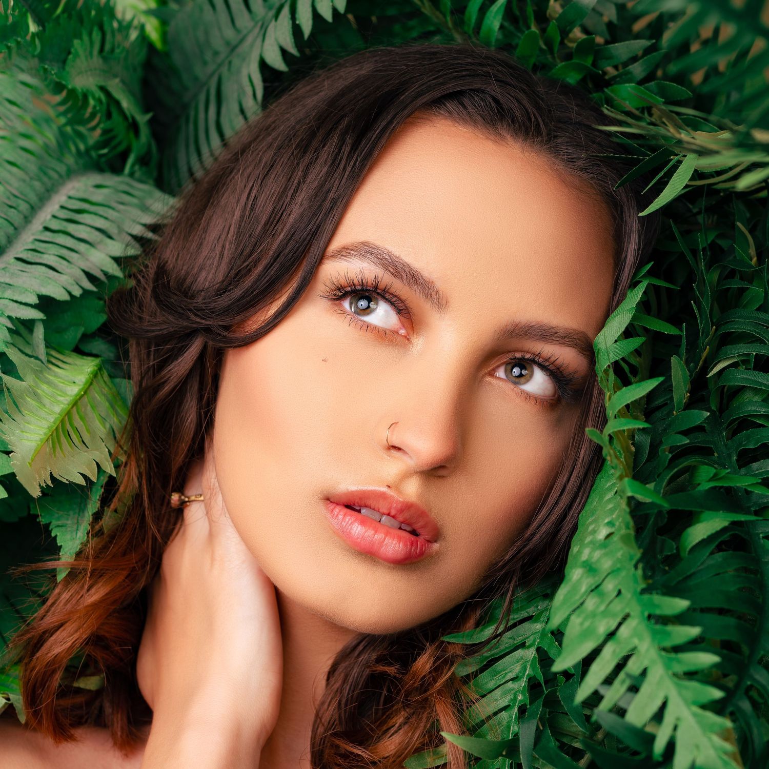 Close-up beauty portrait of a woman with brown hair and light eyes, surrounded by lush green foliage.