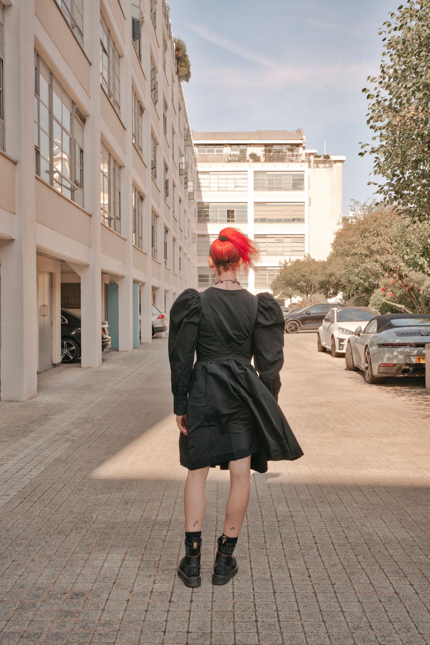 Woman with red hair in black puff-sleeve dress and platform boots stands on urban cobblestone street.