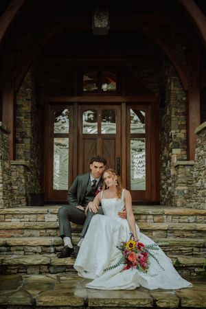 Bride and groom sitting on stone steps of a rustic chapel entrance, bride holding colorful bouquet with sunflowers.