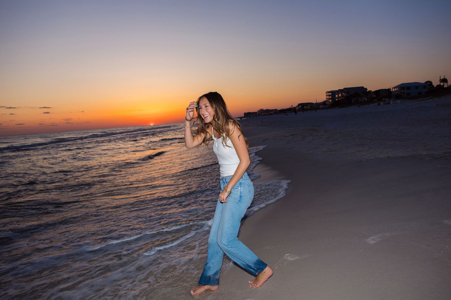 Young woman walking barefoot on a beach at sunset, wearing blue jeans and white top, waves gently touching the shore.