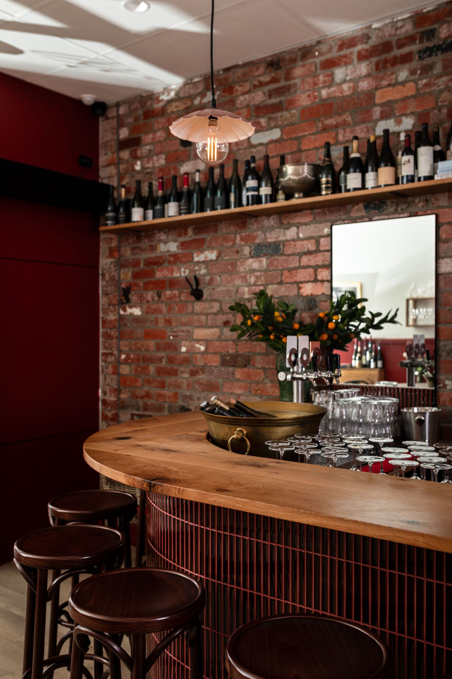 Cozy wine bar interior with a curved timber counter, wooden bar stools, exposed brick wall, pendant light and shelves of wine bottles.