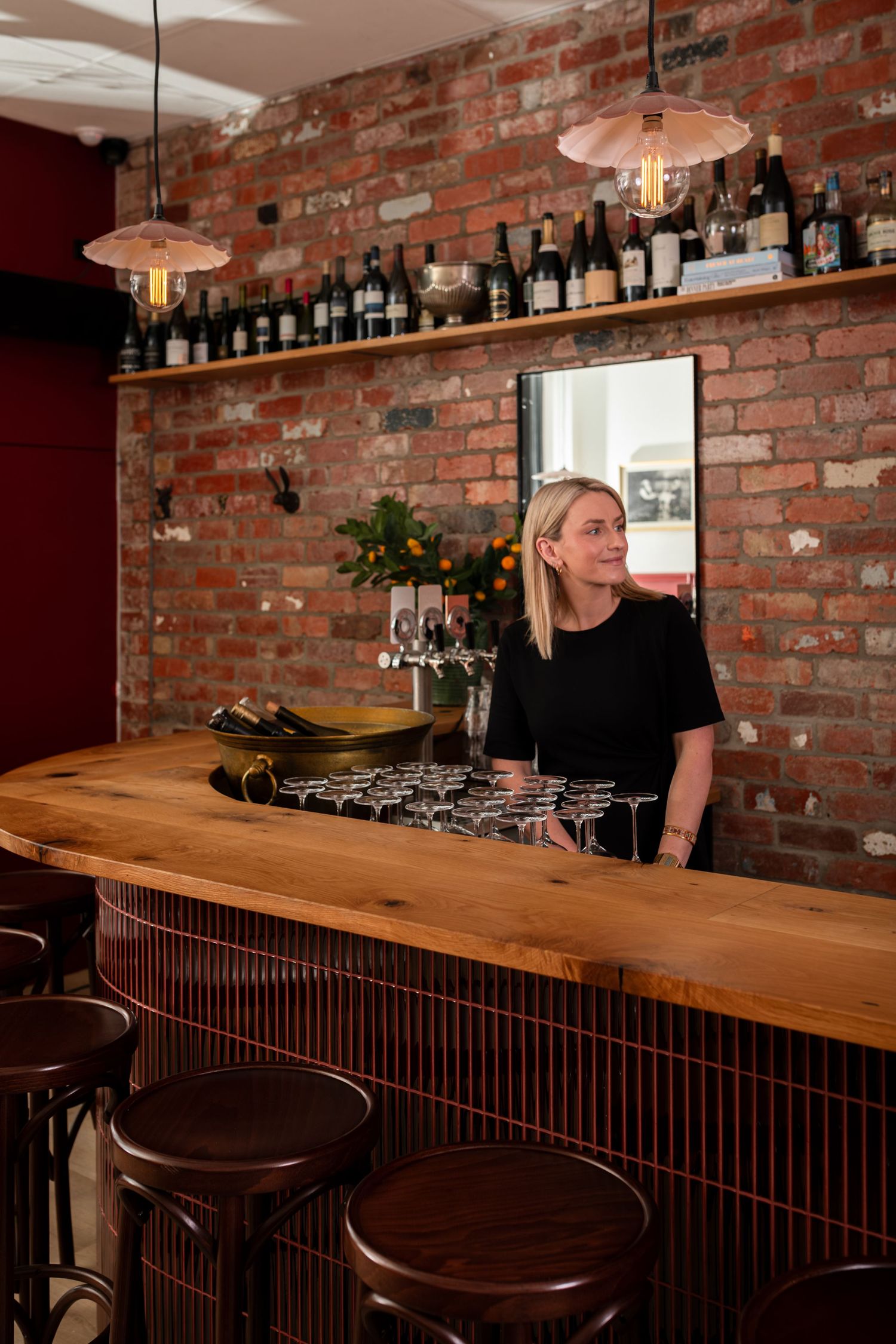 Smiling woman stands behind a rustic wooden bar with champagne glasses, exposed brick walls, wine bottle shelf and pendant lights.