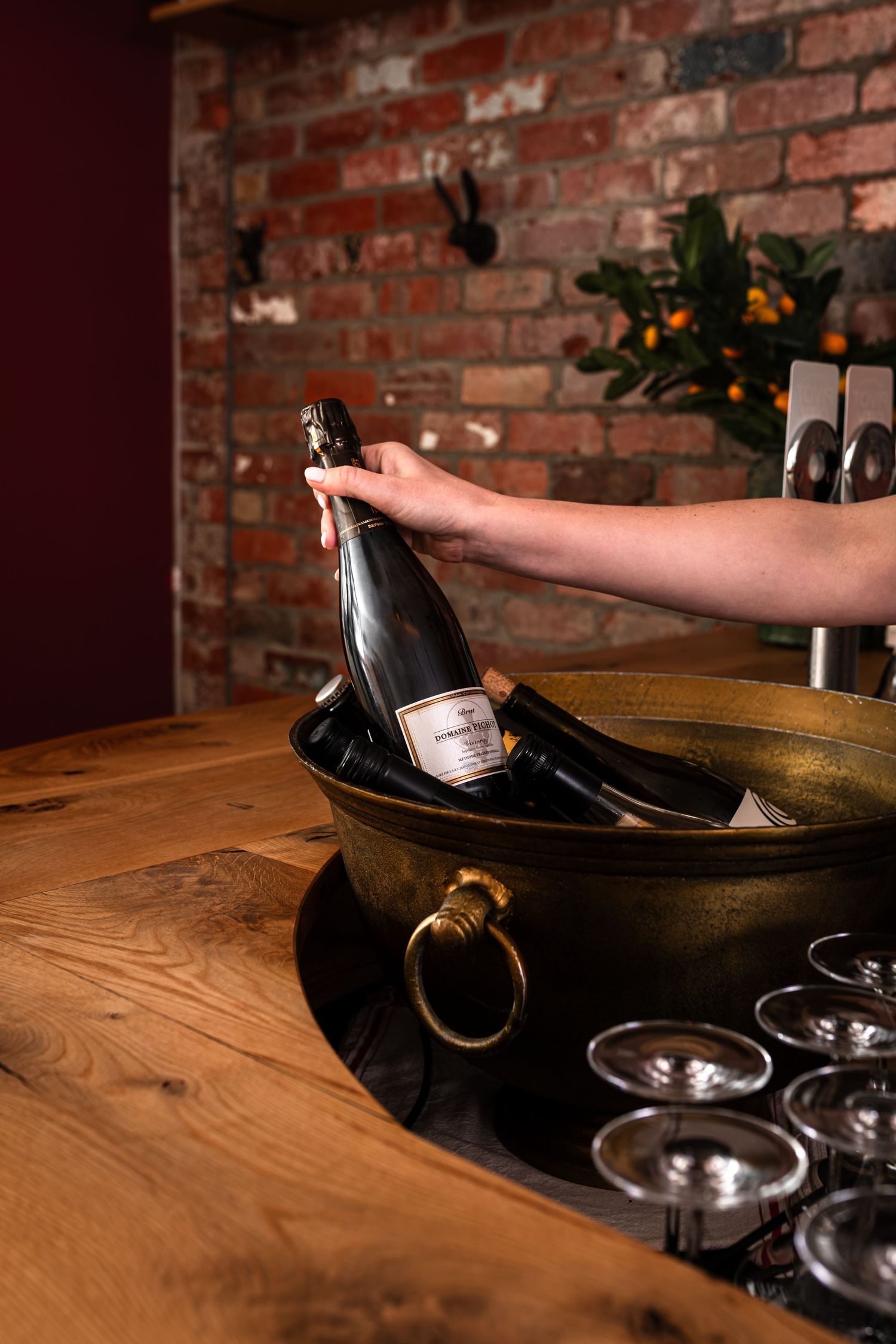 Hand lifting a sparkling wine bottle from a brass ice bucket on a rustic wooden bar with champagne glasses and a brick wall behind.