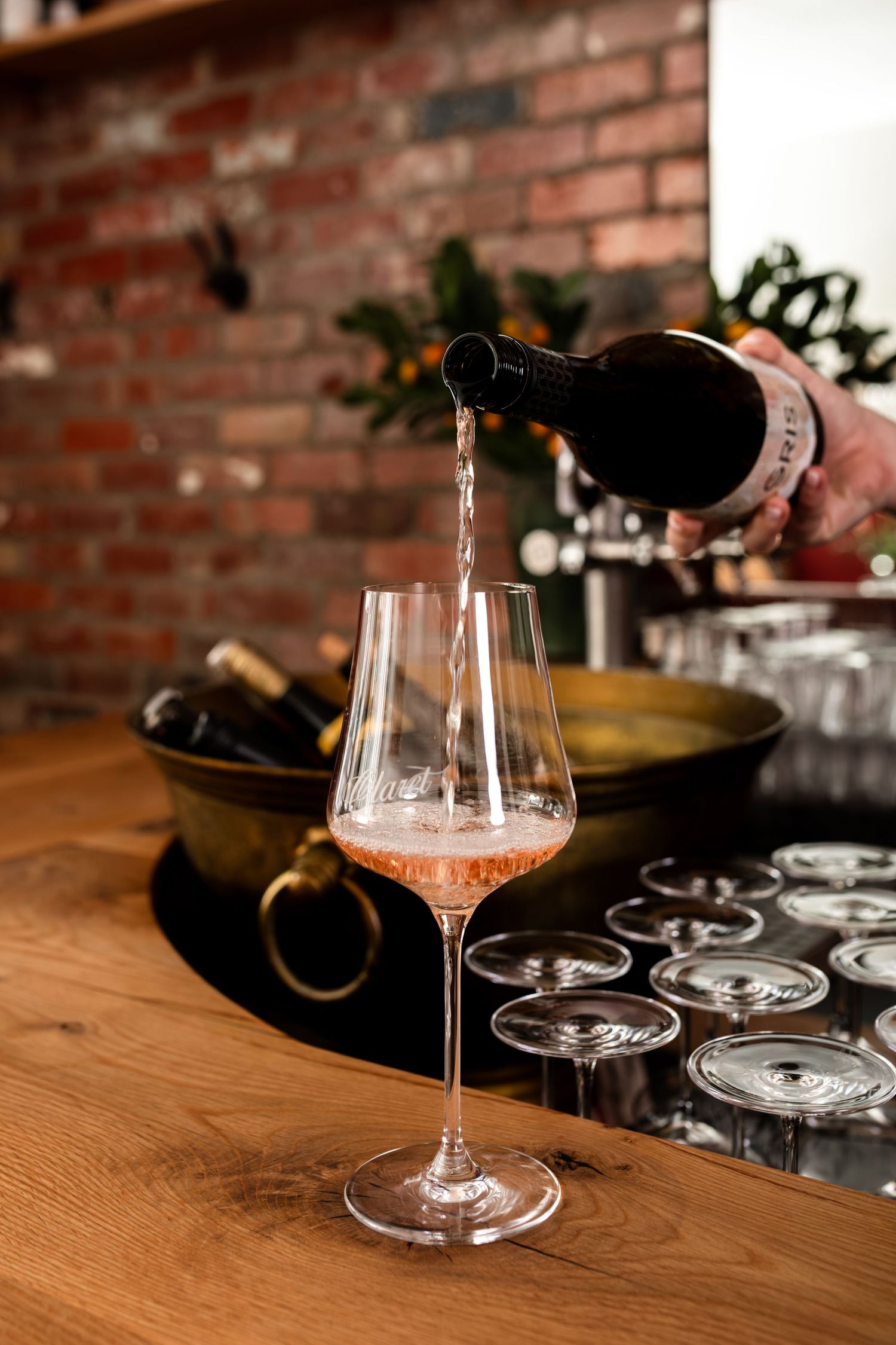 Rosé wine pours from a dark bottle into a stemmed glass on a wooden bar, with wine glasses and a brass ice bucket against a brick wall.