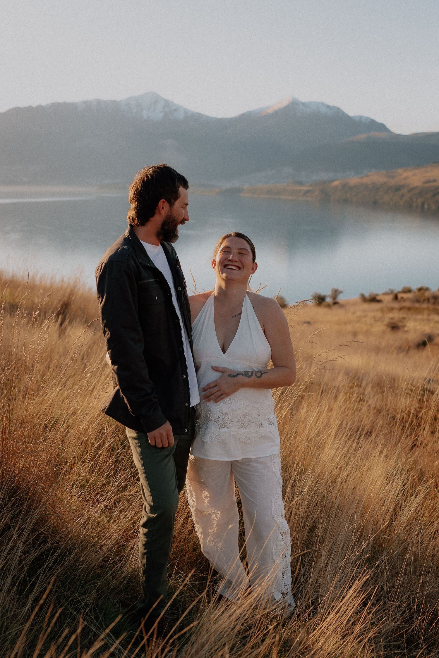 Couple sharing a romantic kiss at golden hour by a serene mountain lake, bride in lace gown, groom in dark suit.