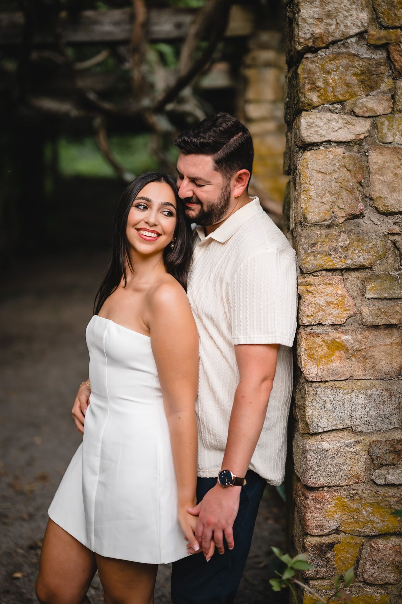Couple posing by stone wall, woman in white mini dress smiling, man in cream shirt holding hands outdoors.