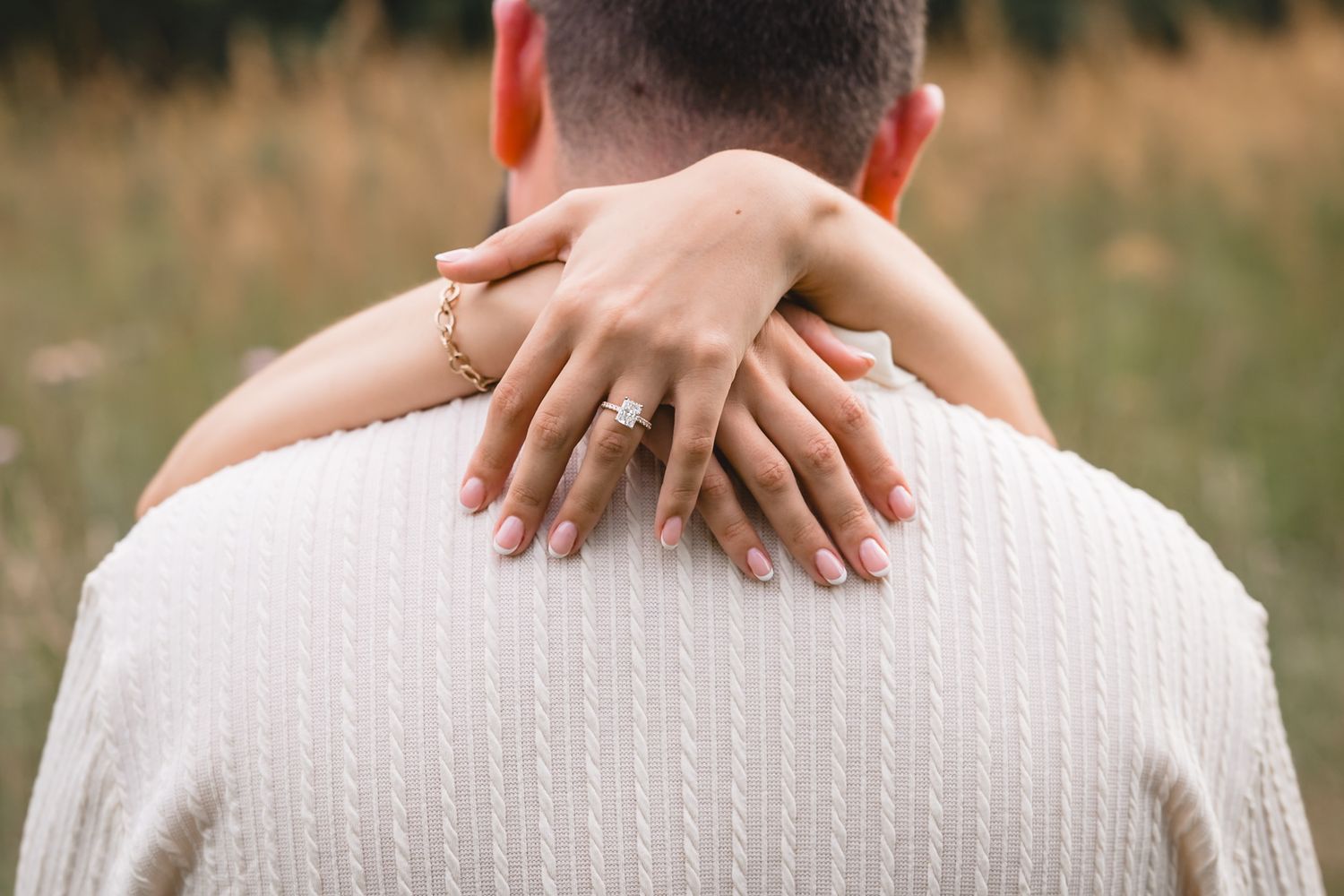 Woman with engagement ring and gold bracelet embraces man from behind in a romantic outdoor field setting.