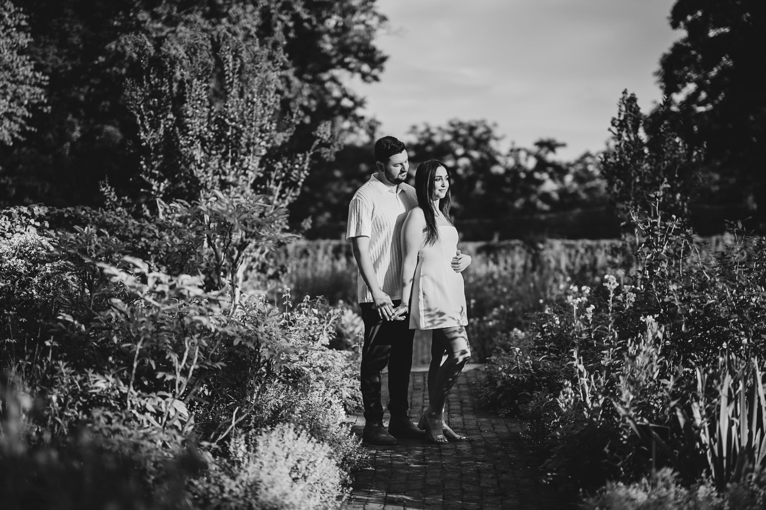 Black and white photo of a couple standing on a brick path surrounded by lush garden greenery.