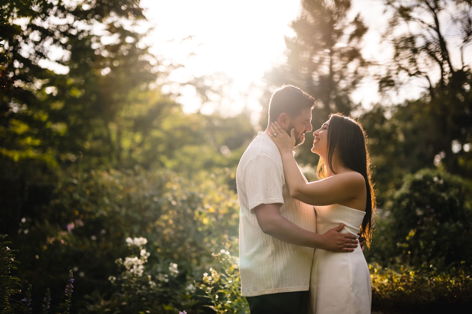 Couple embracing at golden hour in a lush garden, sharing a romantic moment with warm sunlight glowing behind them.