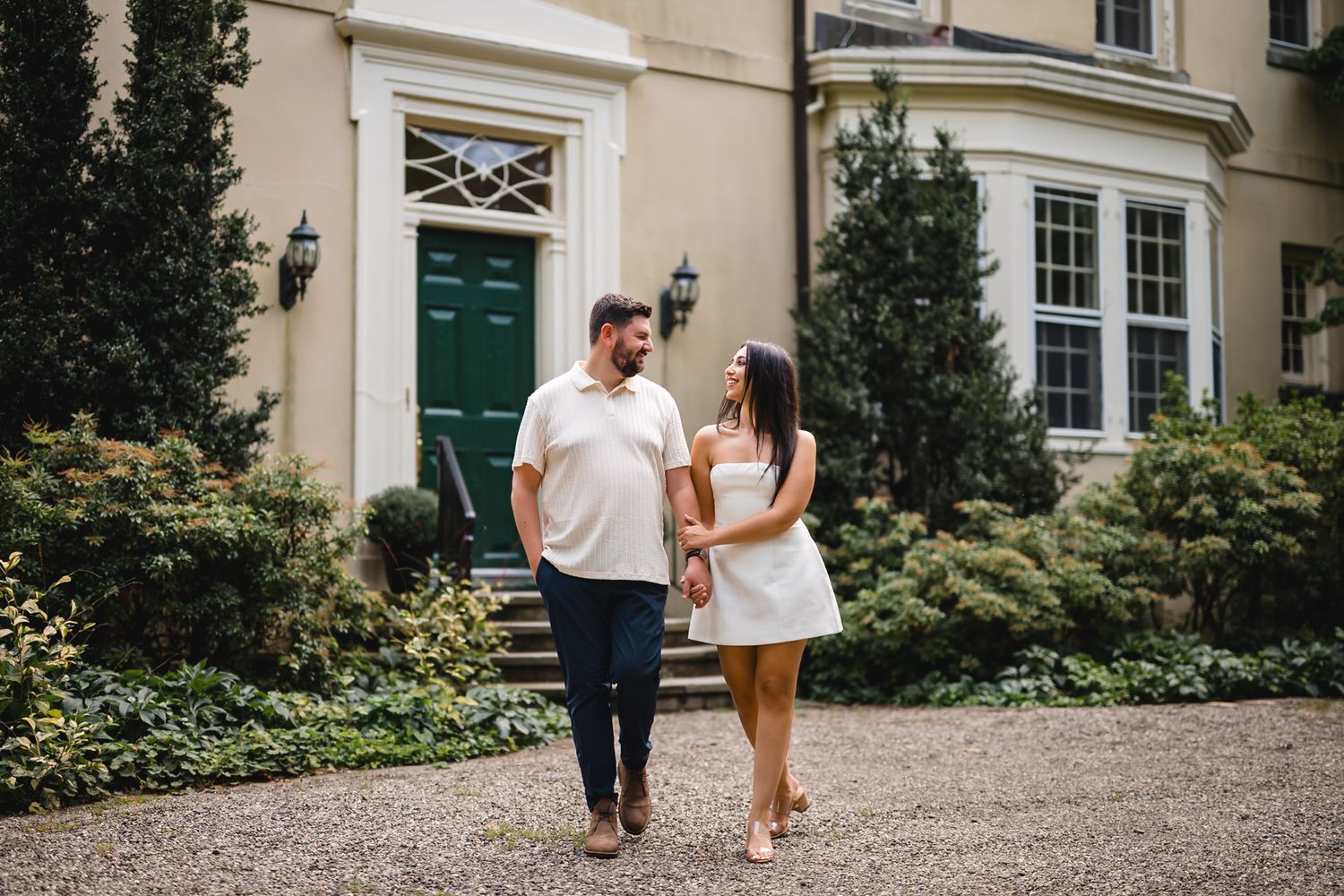 A couple holds hands and walks together outside an elegant estate with lush greenery and a green door.