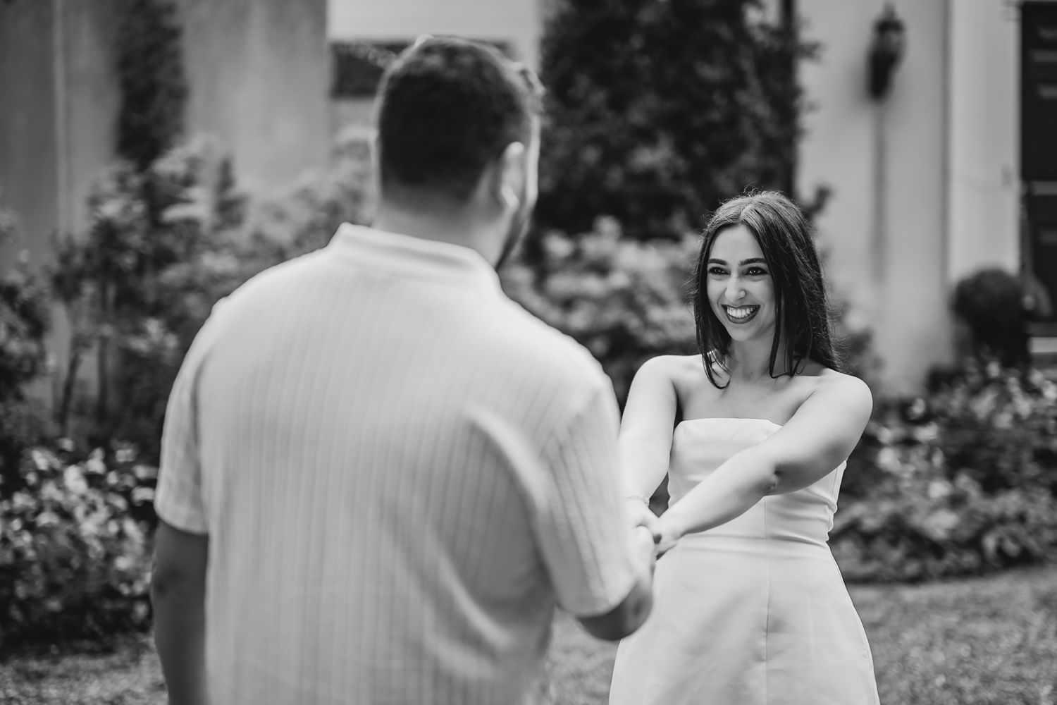 Black and white photo of a smiling woman in a white dress holding hands with a man outdoors.