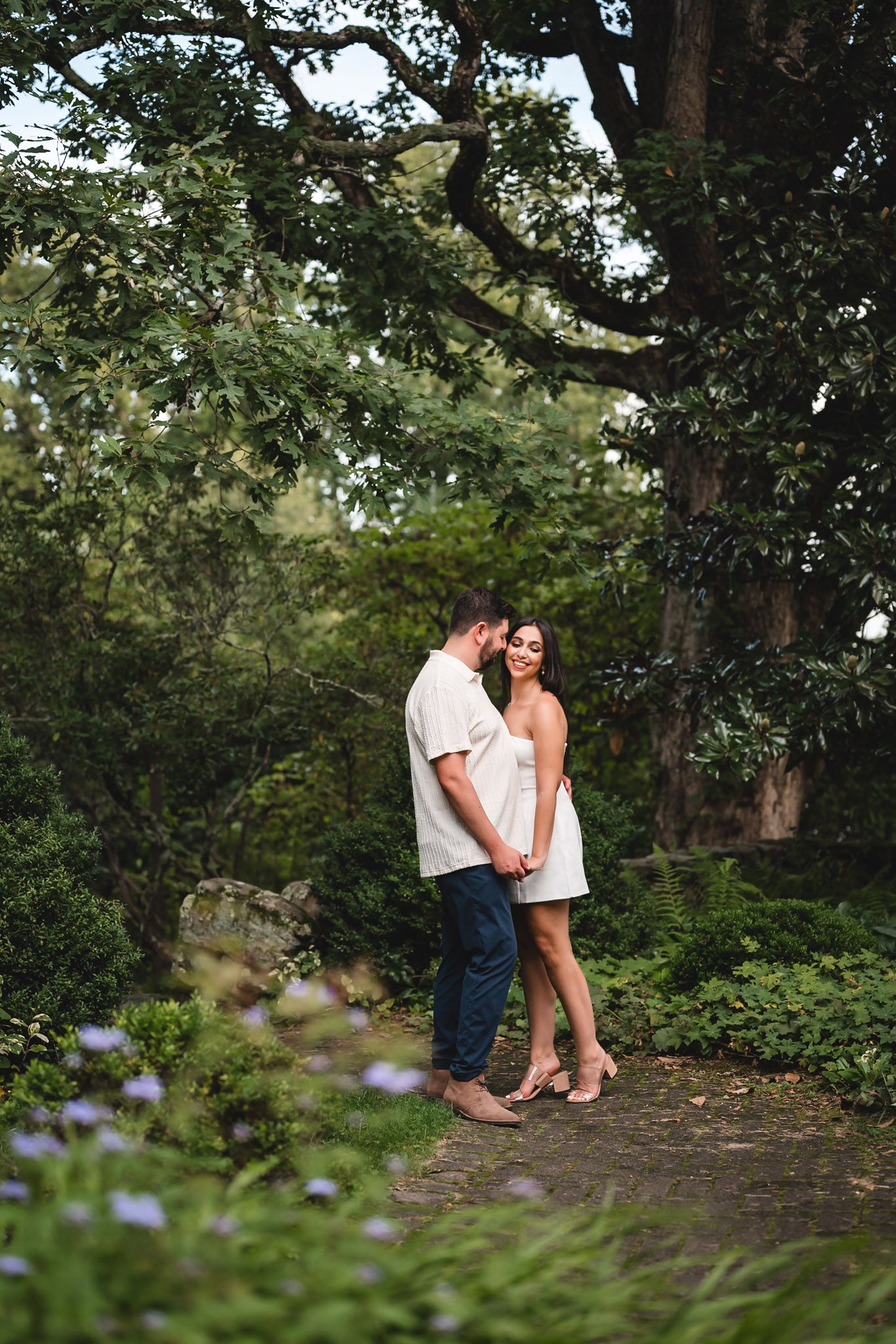 Couple embracing on a garden path surrounded by lush greenery and a large tree, woman in white dress.