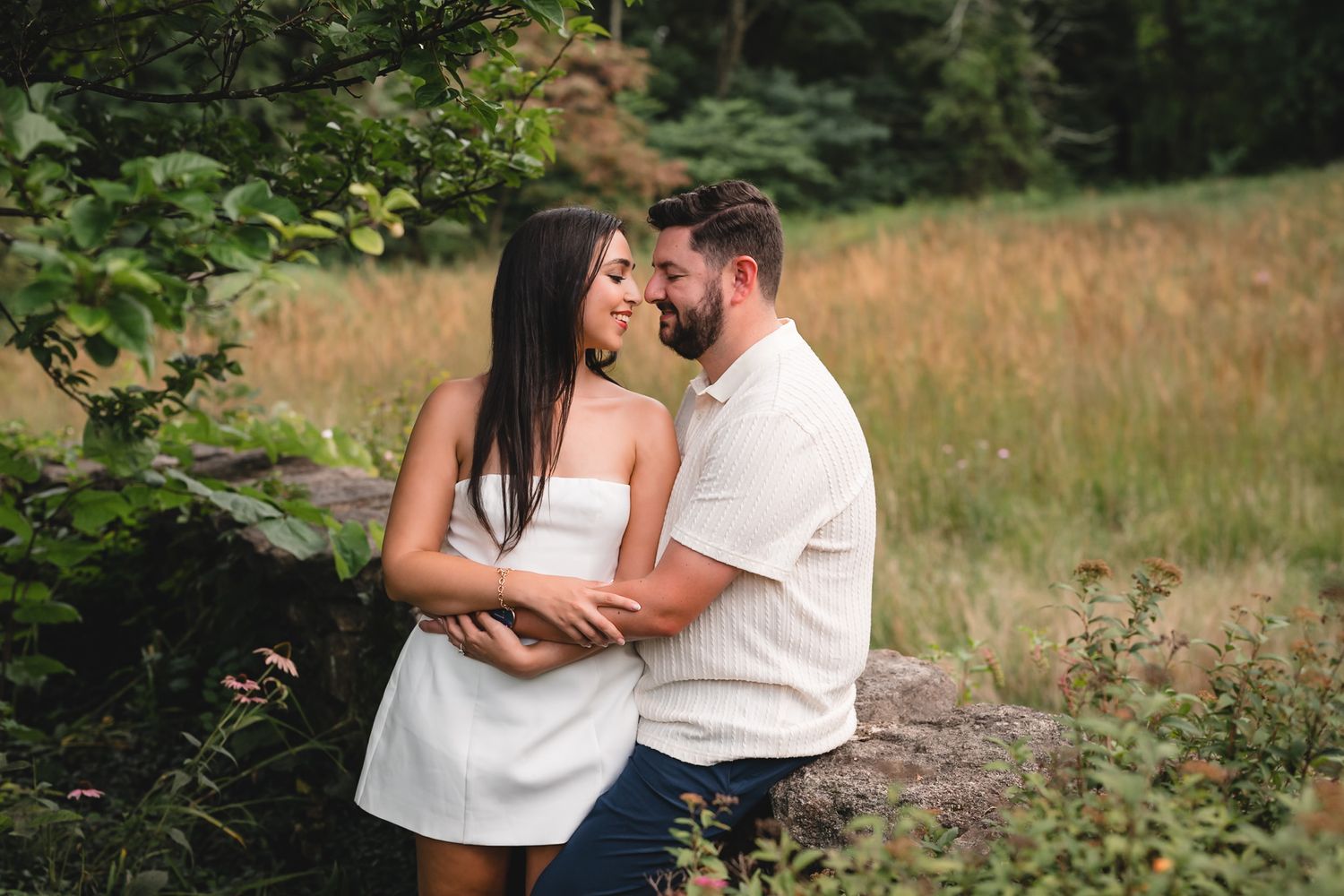A couple in white outfits share a romantic moment surrounded by lush greenery and golden grass in an outdoor setting.