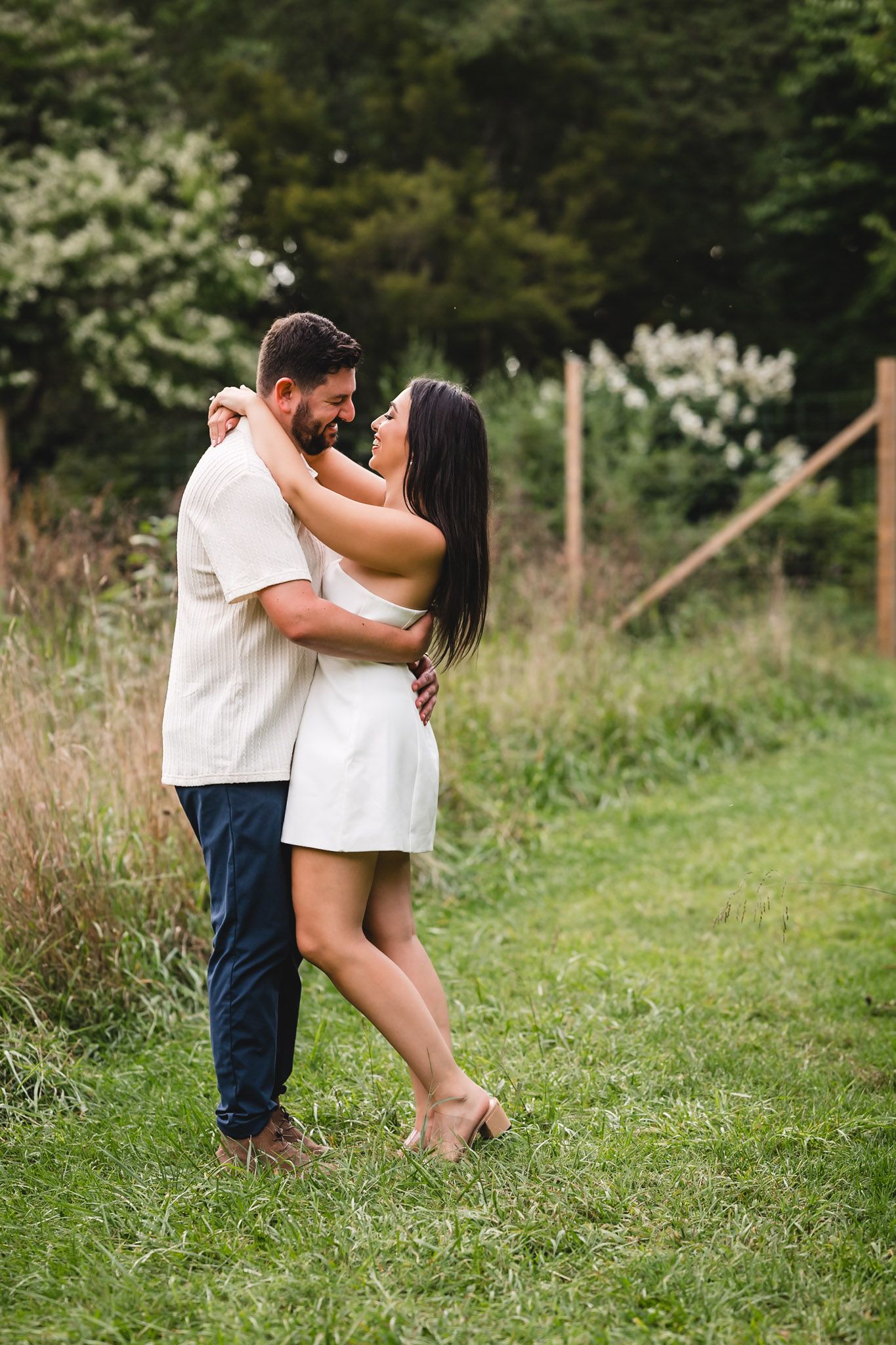 Couple dancing and embracing outdoors on green grass with wildflowers and wooden fence in background.