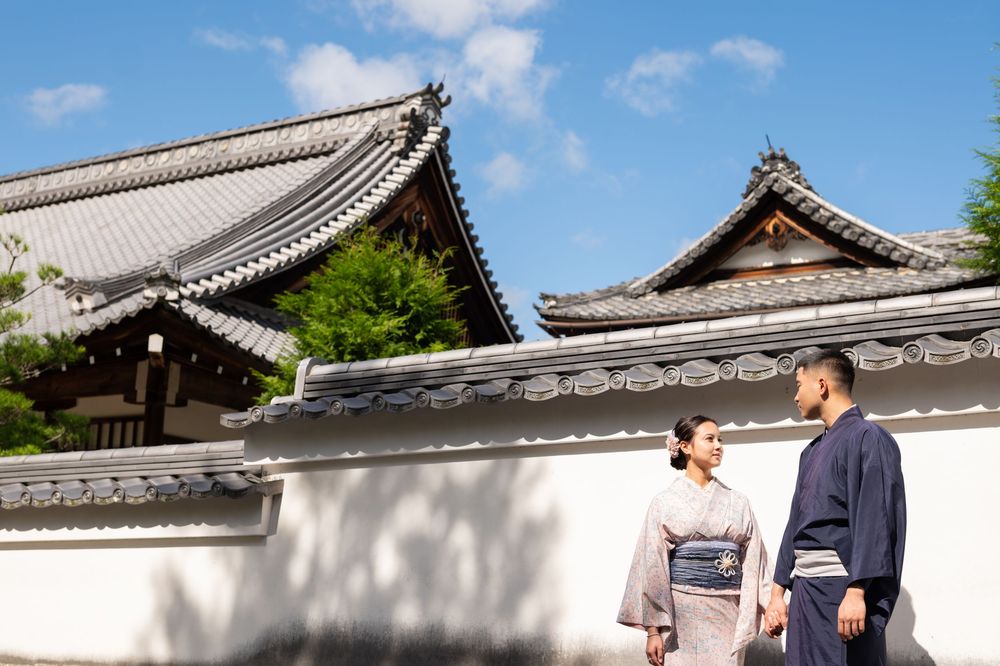 Couple in traditional Japanese kimono strolling past white walls and historic temple rooftops under a blue sky.