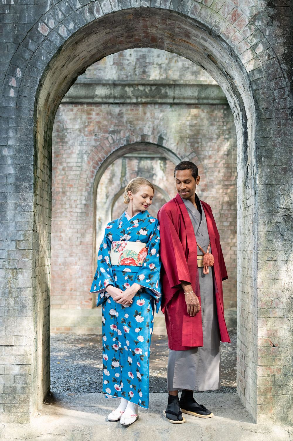 Couple in traditional Japanese kimono and haori standing under stone archway in cultural setting.