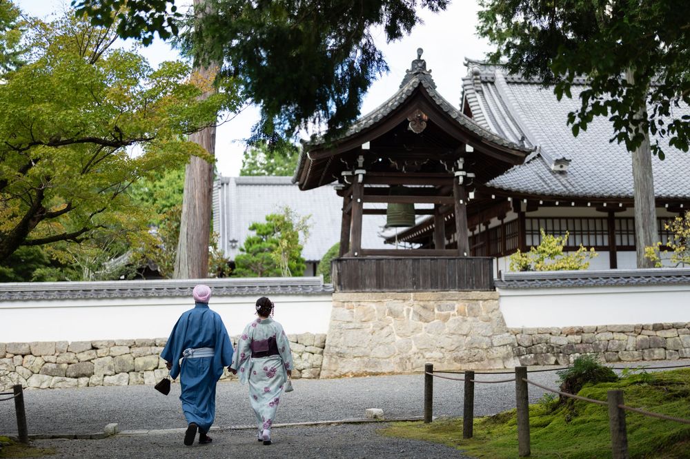 Two people in traditional Japanese kimono walk toward a historic shrine gate surrounded by lush trees and stone walls.