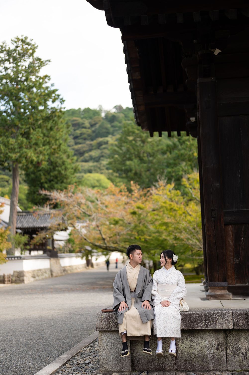 A couple in traditional Japanese kimono sitting together on a stone ledge outside a wooden temple in Kyoto.