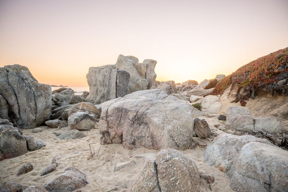 Large granite boulders scattered on sandy beach at sunset with warm orange sky in background.
