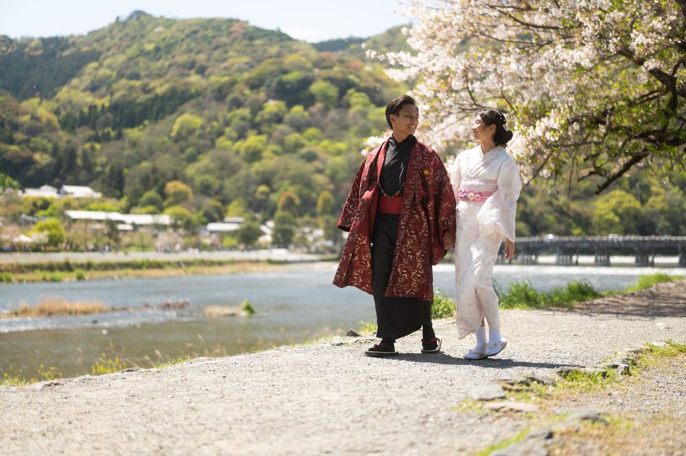 Couple in traditional Japanese kimono walking along a riverside path near cherry blossoms with forested mountains in the background.