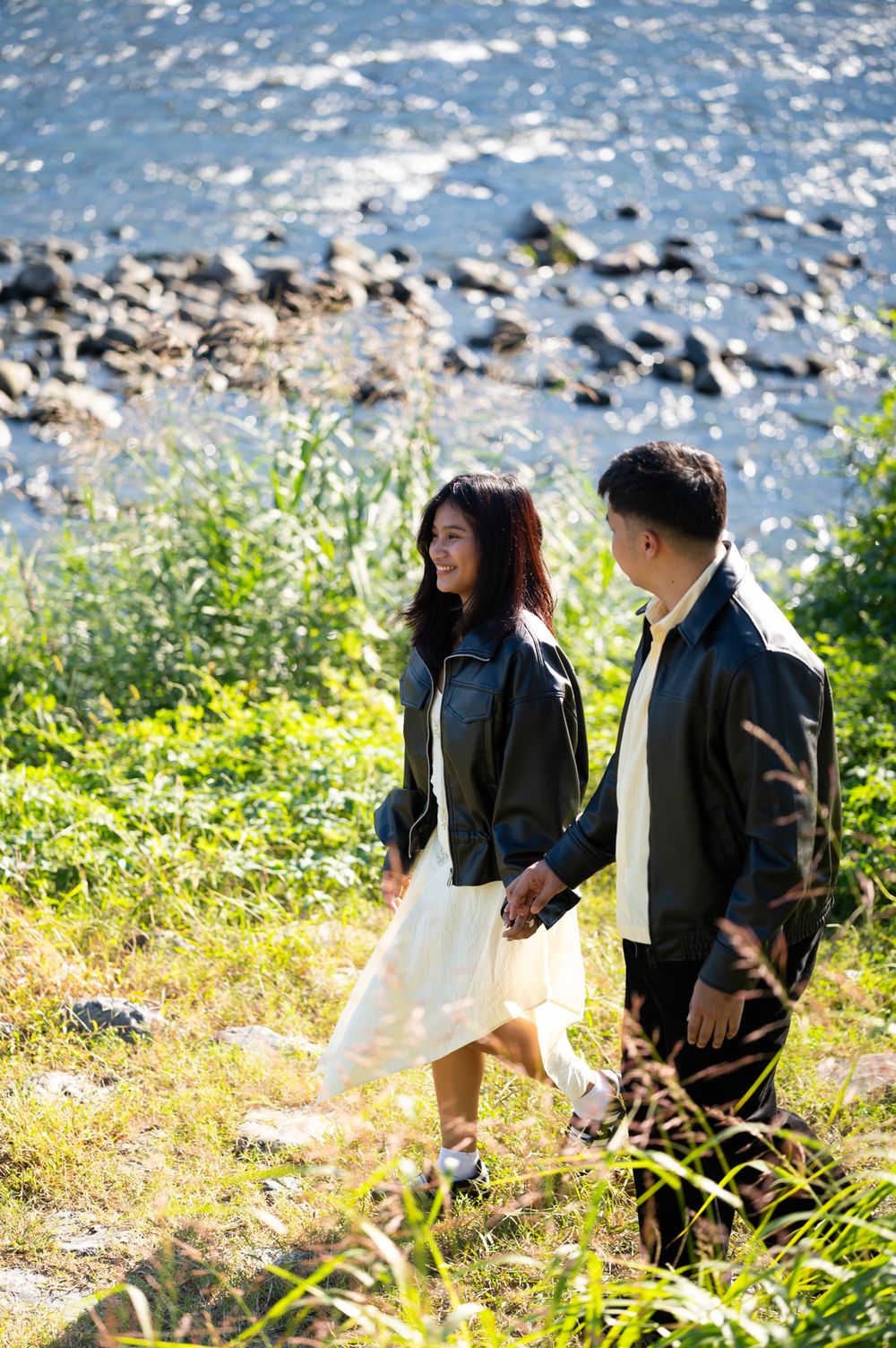 Smiling couple in black leather jackets holding hands and walking through tall grass beside a rocky riverbank on a sunny day.