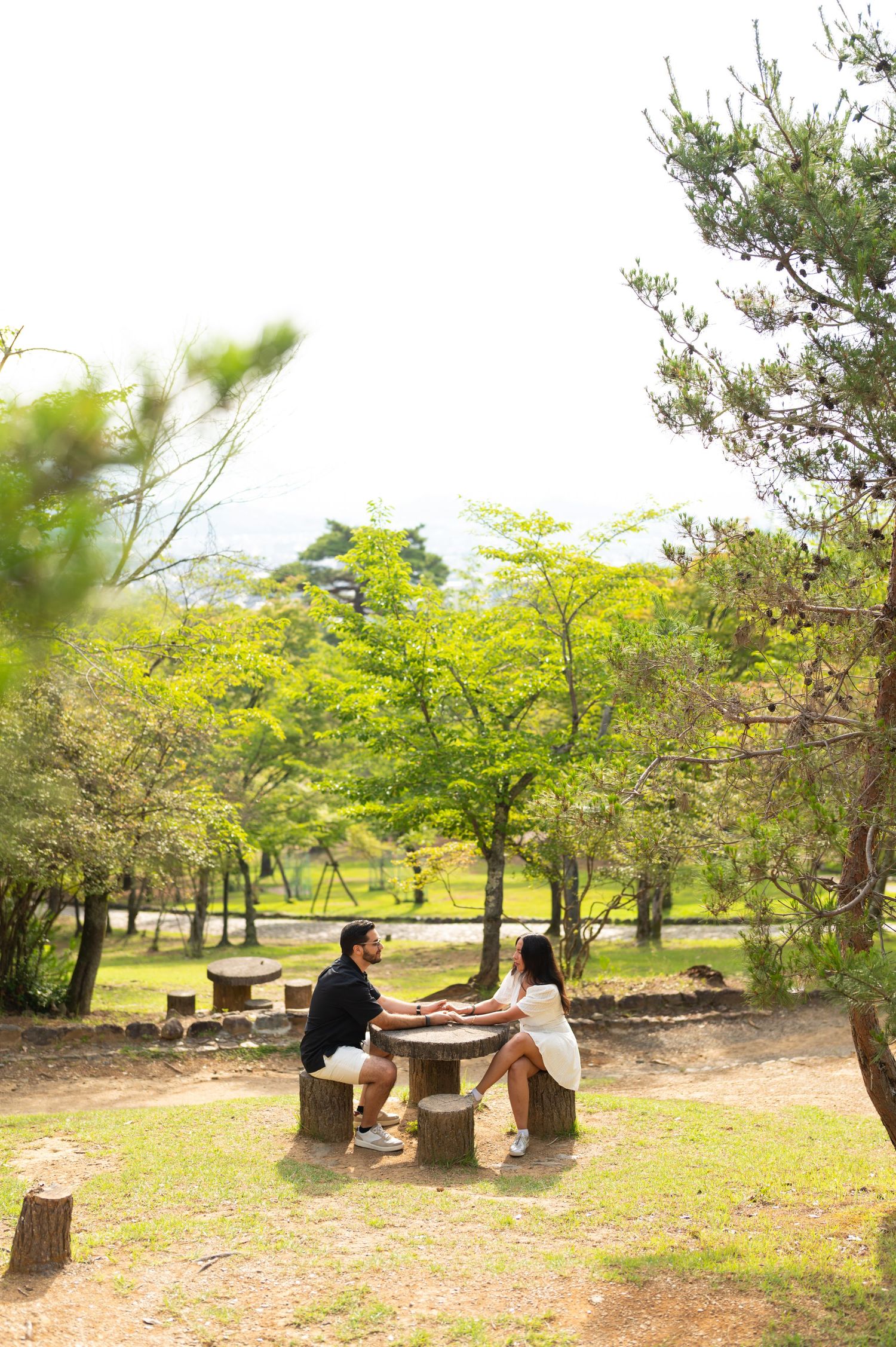 A couple sits holding hands at a wooden stump table in a sunny, tree-filled park with green foliage and a bright sky.