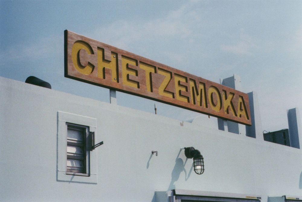 Wooden sign reading CHETZEMOKA in yellow letters mounted on the roof of a white building under a pale blue sky.
