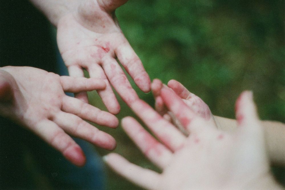 Several outstretched hands with red berry-stained fingers and palms reach toward each other against a blurred green background.