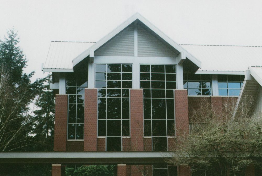 Modern brick building with a steep gabled roof and tall glass curtain wall windows, framed by evergreen trees under a cloudy sky.