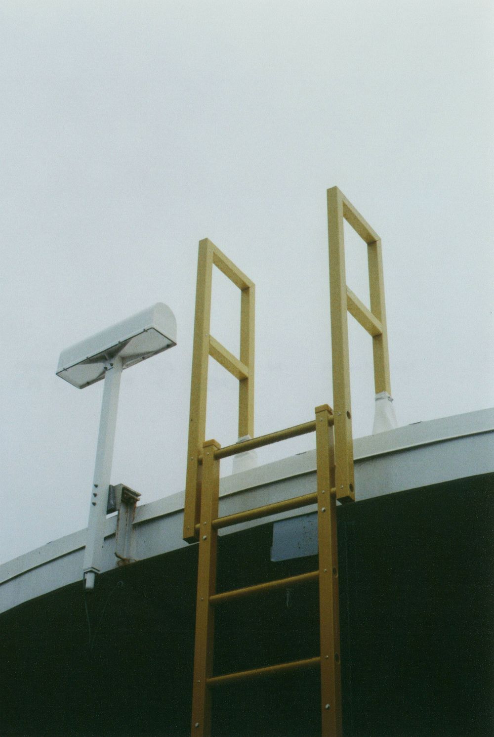 Yellow fiberglass access ladder with safety cage extending above the rim of a dark storage tank, next to a mounted light fixture.