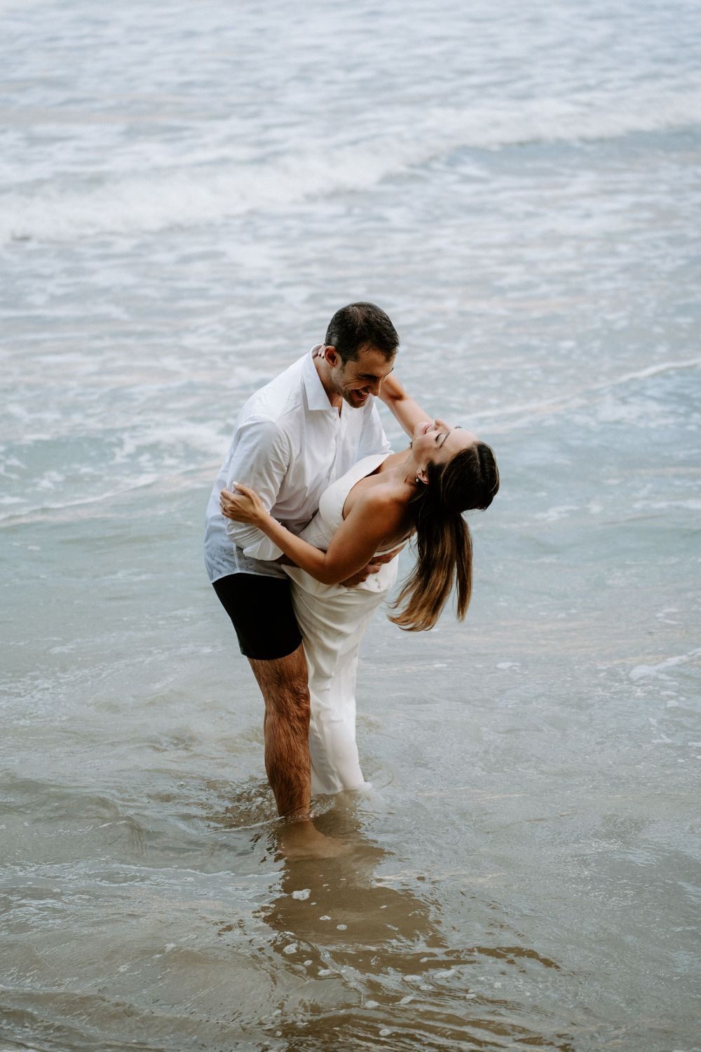 Smiling couple in white outfits dance and dip playfully in shallow ocean water during a romantic beach engagement photo session.