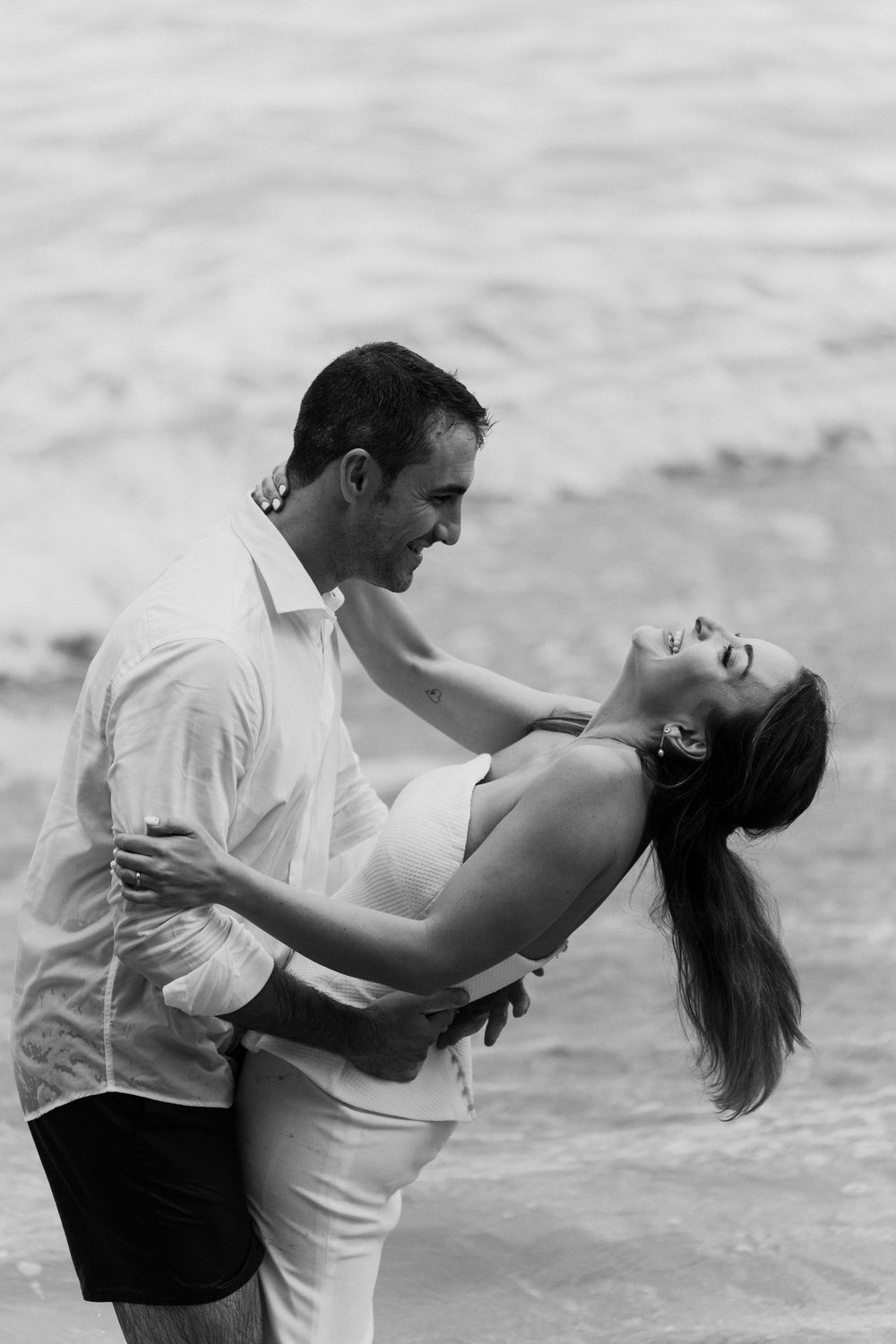 Black and white photo of a smiling couple embracing in a romantic dip beside the ocean waves.