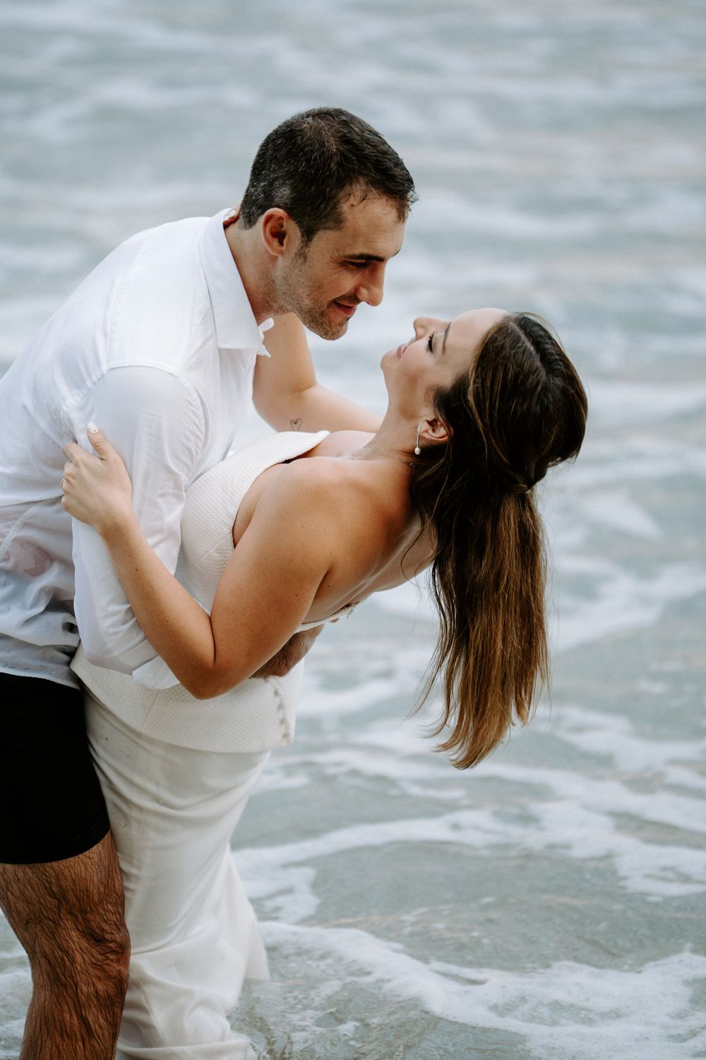 Romantic couple in white outfits embracing and smiling at each other while standing in shallow ocean waves at the beach.