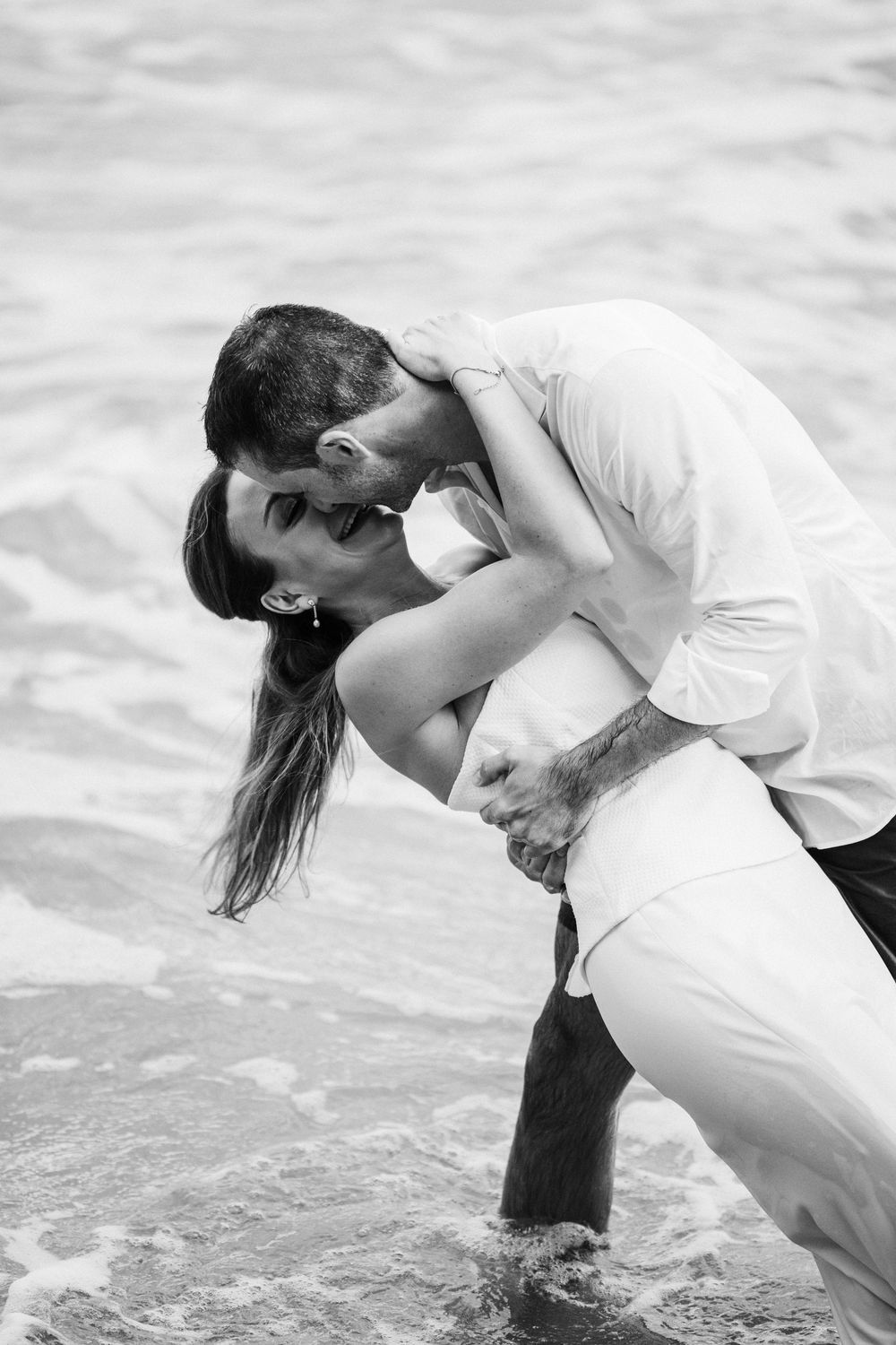 Black and white photo of a couple kissing passionately as the groom dips the bride while standing in shallow ocean waves.