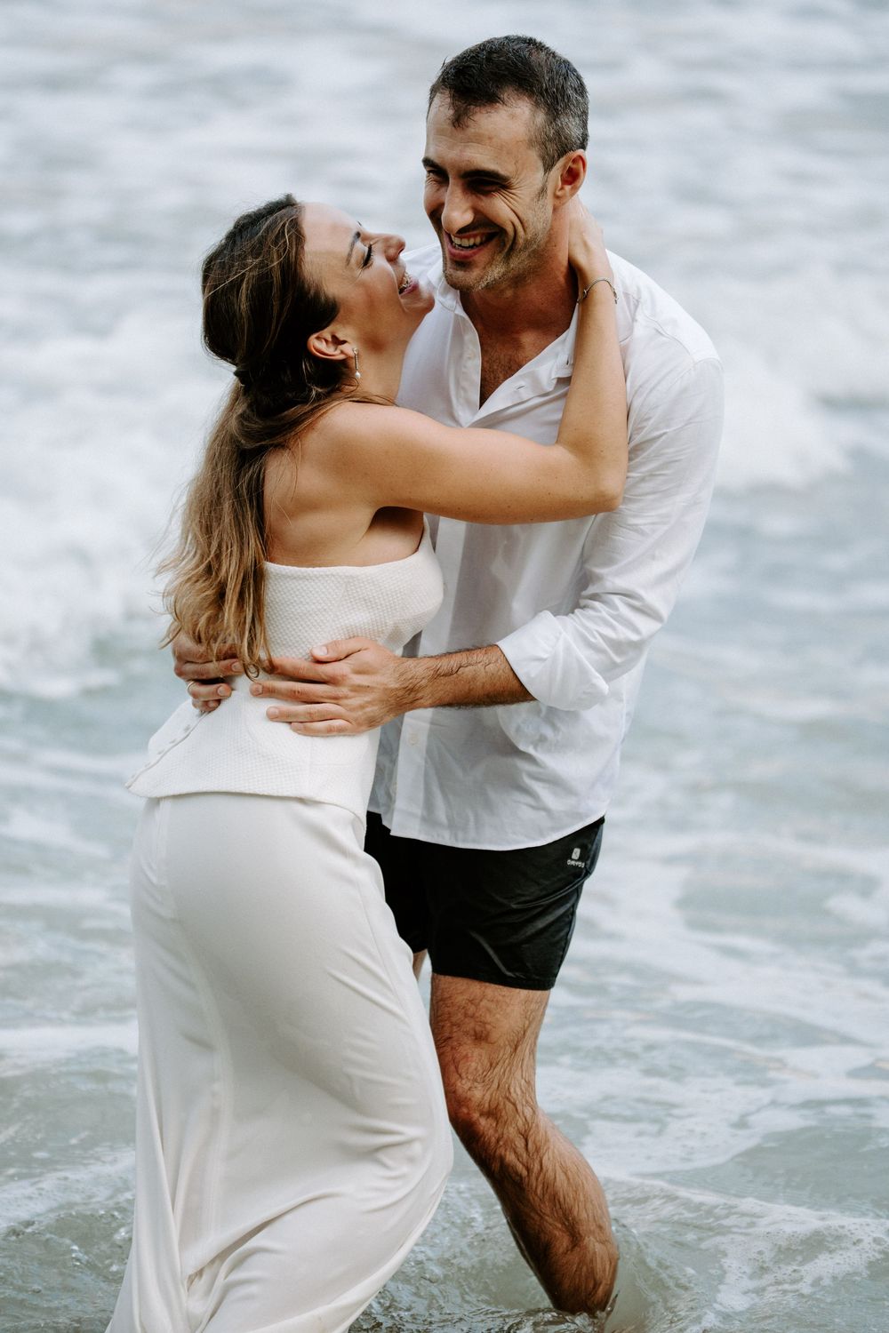 Laughing couple in white outfits hugging in shallow ocean water during a beach engagement photo session.