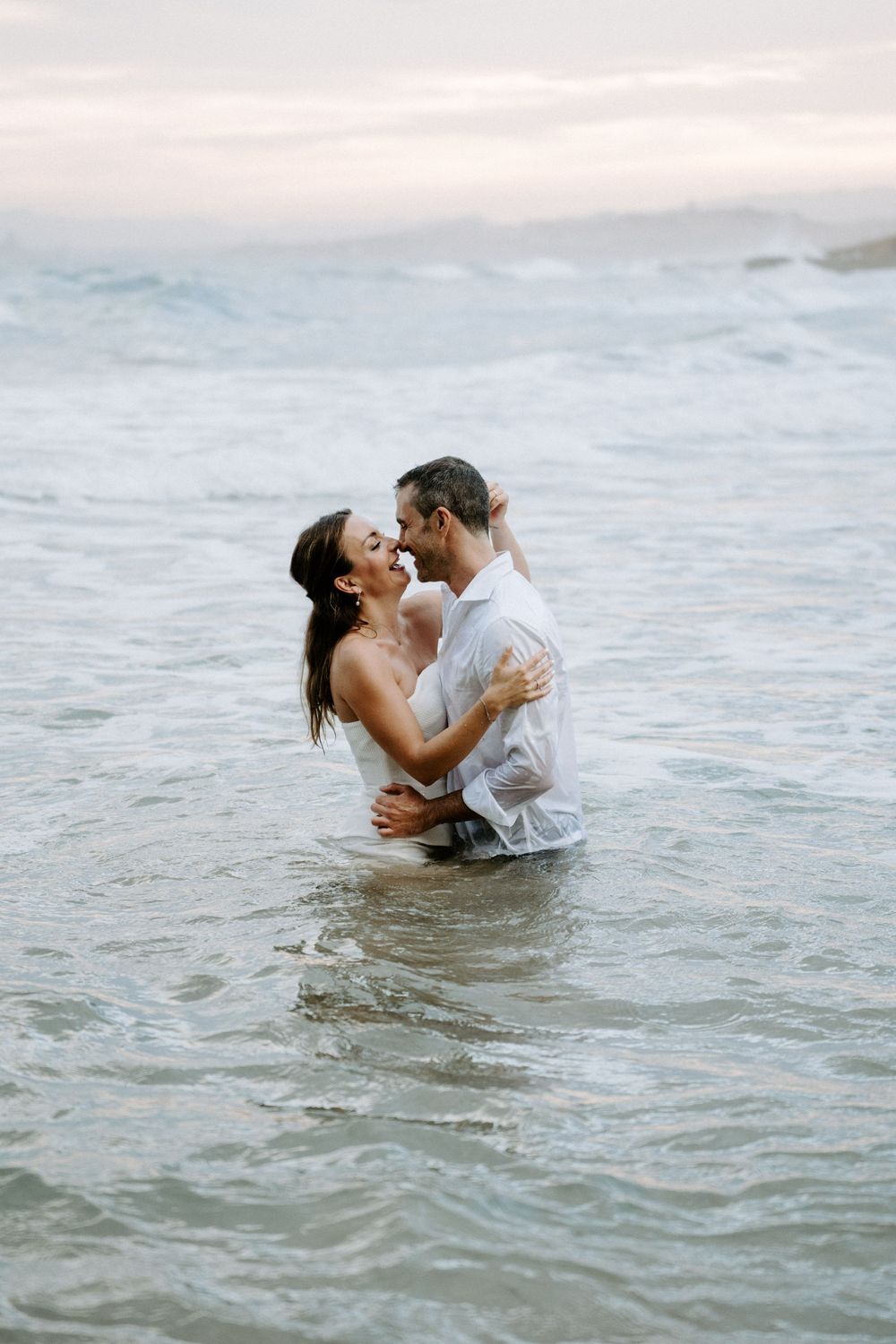 Couple in wet white clothing share an intimate embrace while standing waist-deep in the ocean at sunset.