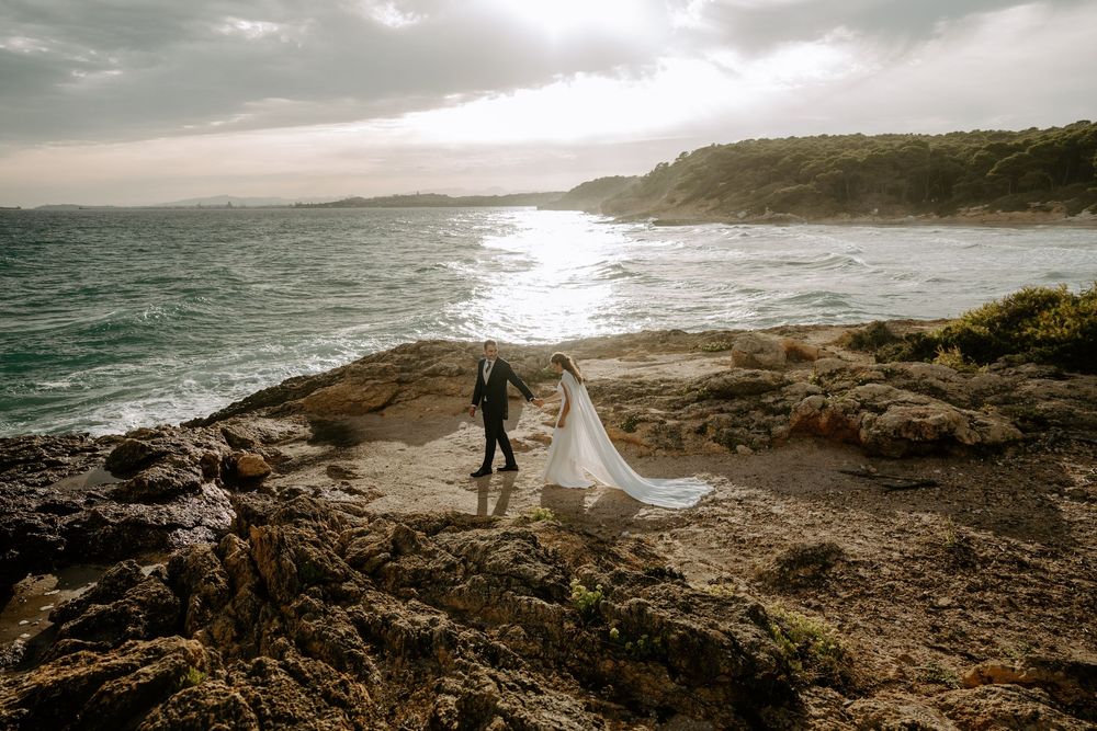 Bride in a flowing white gown and groom in a dark suit walk hand in hand across rocky coastal cliffs overlooking the sea at sunset.
