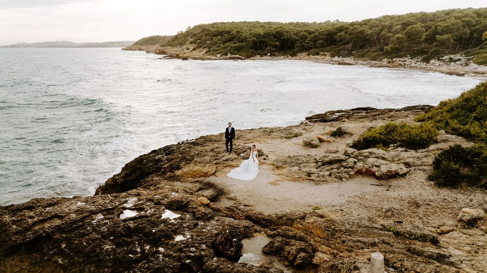 Aerial view of a bride and groom standing on a rocky coastal shoreline beside calm sea waters and green hillside vegetation.