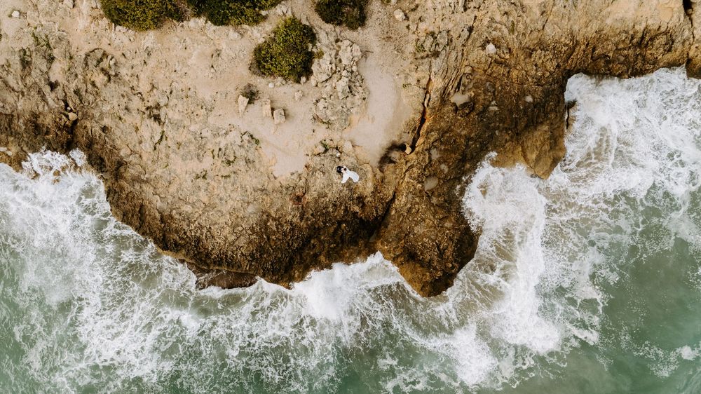 Aerial view of turquoise ocean waves crashing against a rugged rocky coastline with patches of green vegetation.