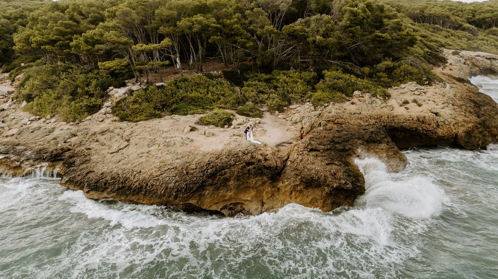 Aerial view of rocky coastline with waves crashing against weathered cliffs and dense green pine trees above the sea.