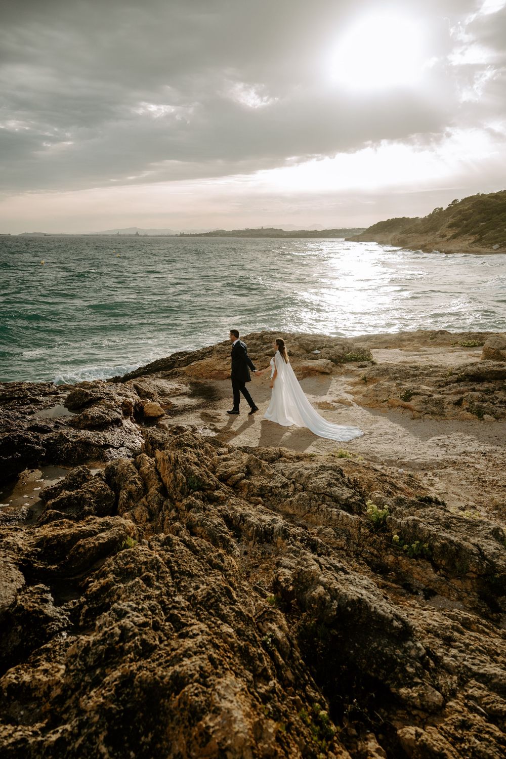 Bride in a flowing white gown and groom in a dark suit walk hand in hand across rocky coastline under a dramatic sunset sky.
